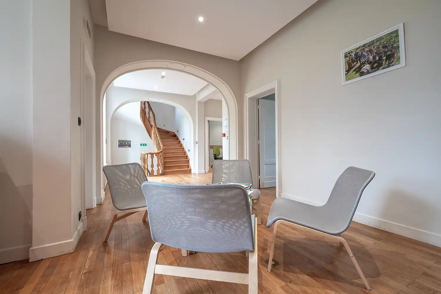 ONIRYX-Manoir-11 Quatre chaises grises entourent une table basse dans une pièce avec parquet, devant un escalier en bois sous une arche.