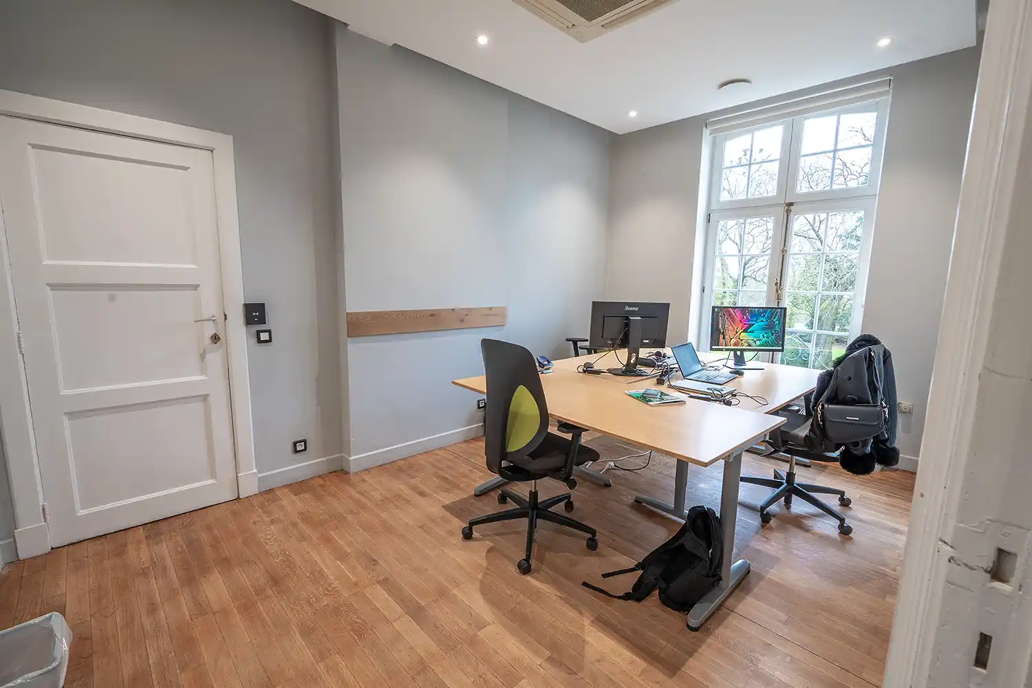 Bureau lumineux avec deux chaises et un bureau en bois clair, équipé d'ordinateurs, face à une grande fenêtre.