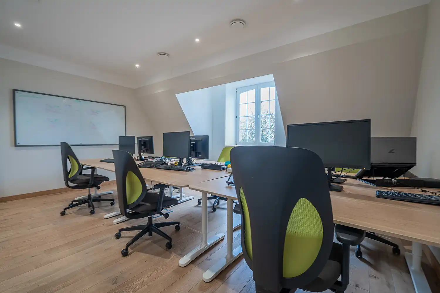 Salle de bureau moderne avec des chaises noires et vertes, plusieurs écrans d'ordinateur sur des tables en bois clair, et une fenêtre lumineuse.
