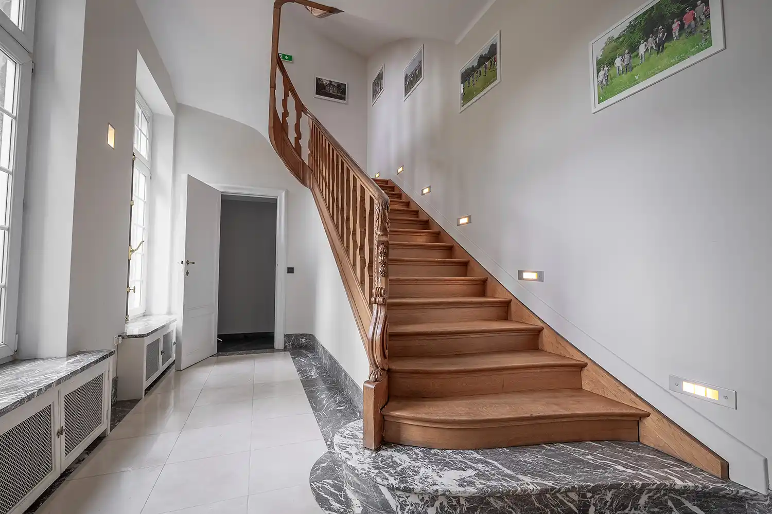 Escalier en bois avec rampe sculptée dans un hall lumineux, sol en marbre et murs ornés de cadres.