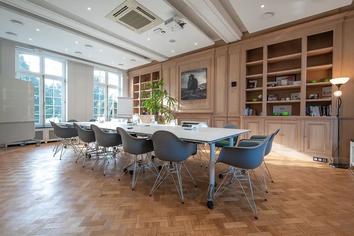 Salle de réunion lumineuse avec table blanche, chaises grises, parquet en bois, bibliothèque intégrée et grandes fenêtres.