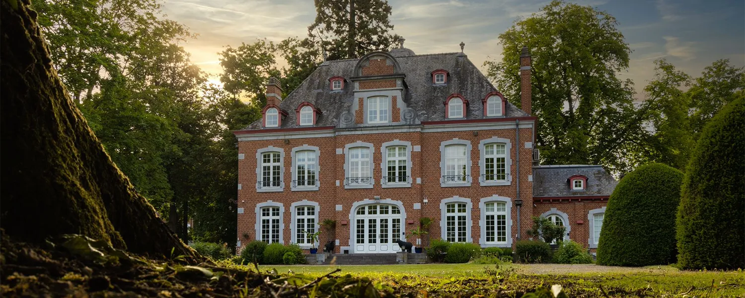 Façade d'une grande maison en briques rouges entourée d'arbres et de buissons, sous un ciel partiellement nuageux.