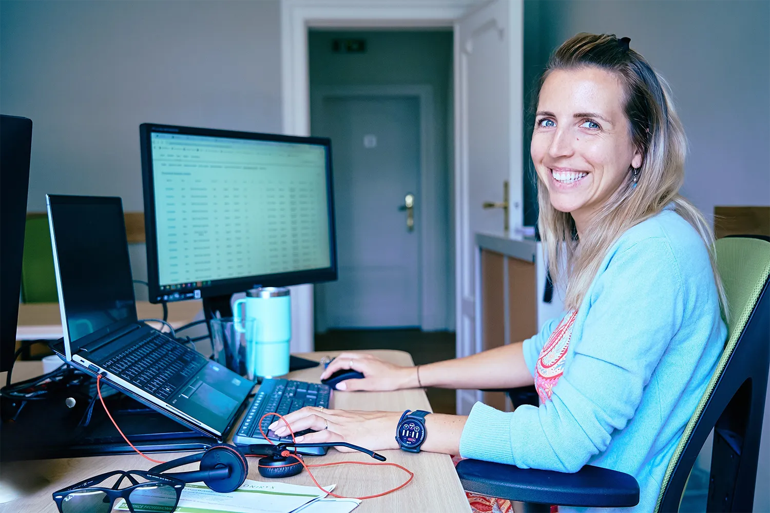 Une femme souriante travaille sur un ordinateur portable avec un écran externe, des écouteurs et une tasse bleue posés sur le bureau.
