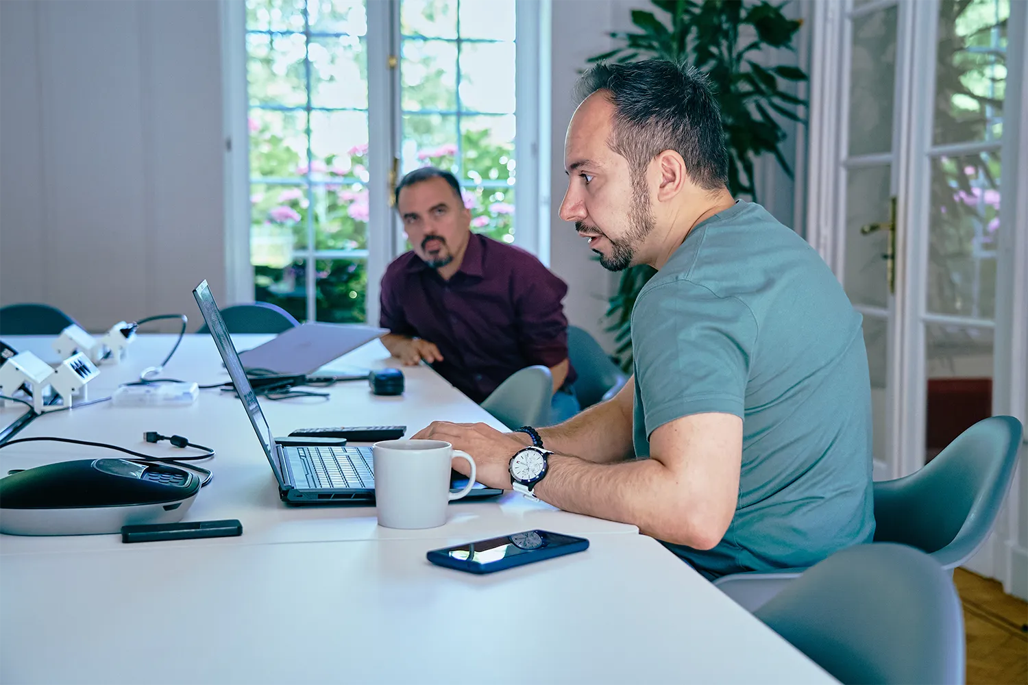 Deux hommes discutent à une table de réunion, l'un utilisant un ordinateur portable, avec une tasse et un smartphone devant lui.