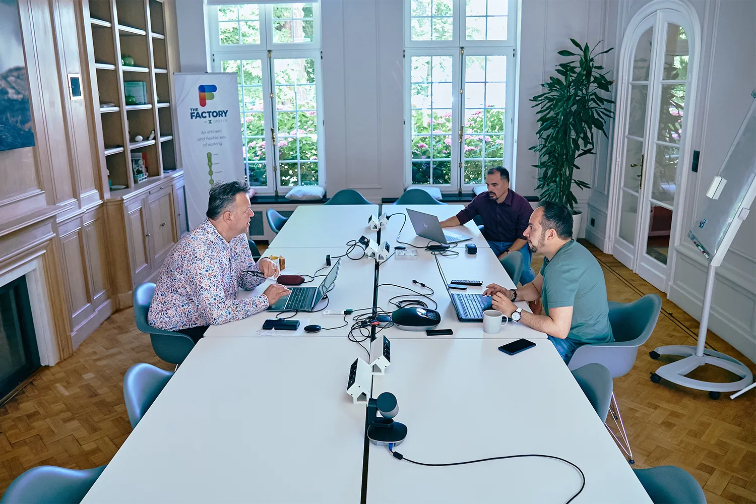 Trois hommes travaillent sur des ordinateurs portables autour d'une grande table blanche dans une salle lumineuse avec des fenêtres et des étagères en bois.
