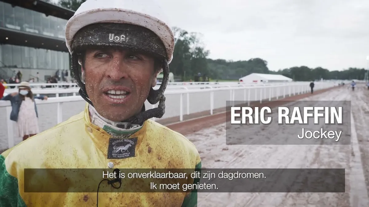 Jockey en tenue jaune et verte, casque blanc, debout sur une piste hippique boueuse avec des spectateurs à l'arrière-plan.